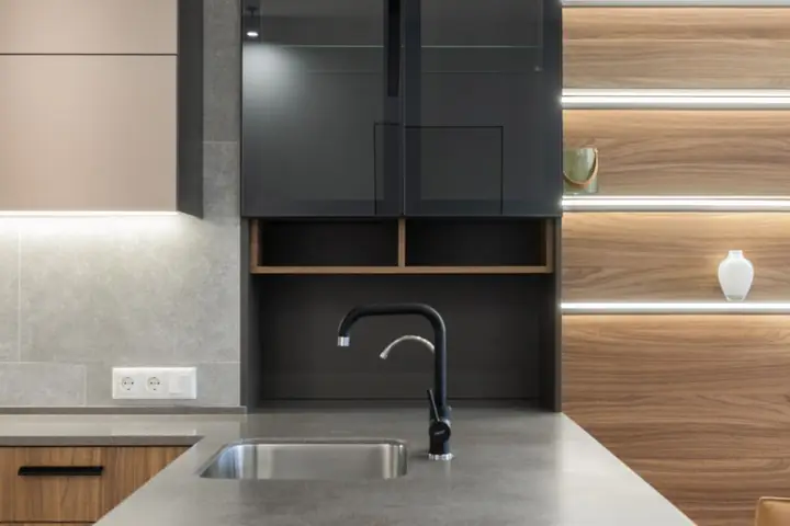 Gray kitchen counter with faucet and sink near cabinets and light decorations on wooden wall in modern apartment at home