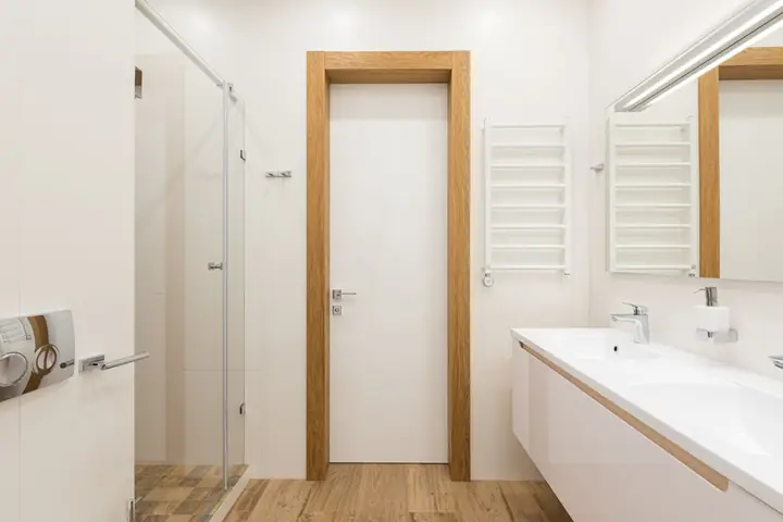 White door and furniture in modern bathroom with glass shower cabin and wooden elements reflecting in mirror
