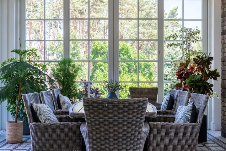 Indoor dining space with wicker chairs and lush potted plants by large windows