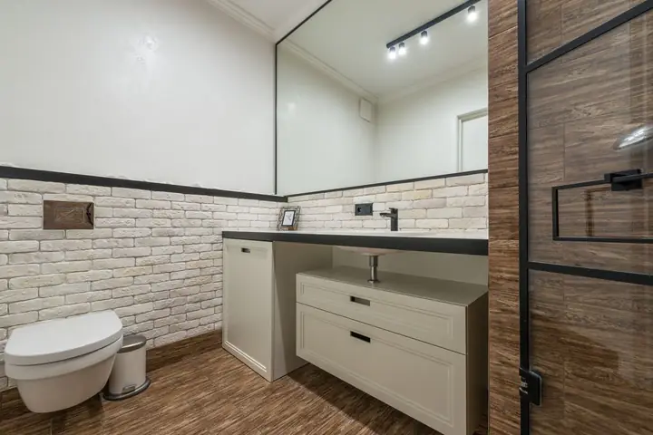 Toilet placed at wall near white cabinet and sink with mirror in bathroom with glass shower cabin in apartment